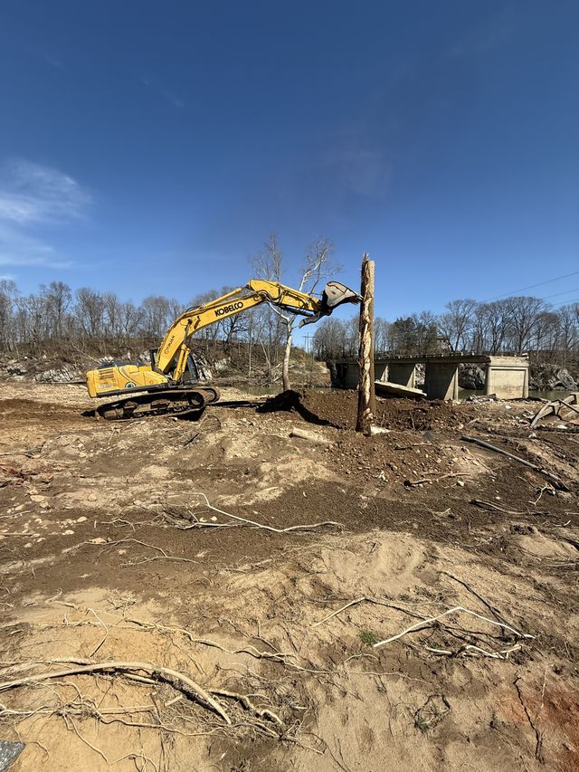 Excavator working near the broken concrete remains of a damaged bridge crossing a creek during debris removal, western North Carolina