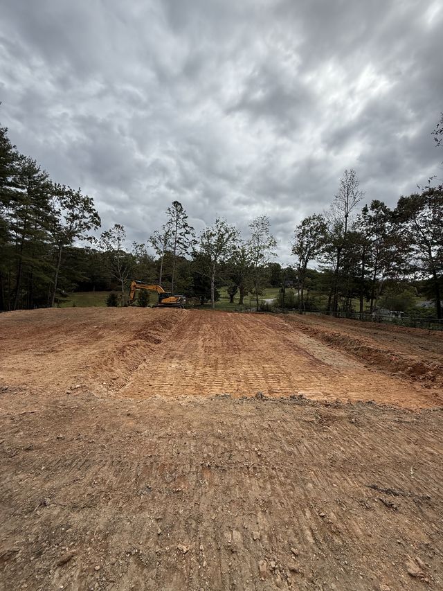 Finish-graded building pad with scarified soil in tidy rows under a dramatic cloudy sky, excavator in the distance