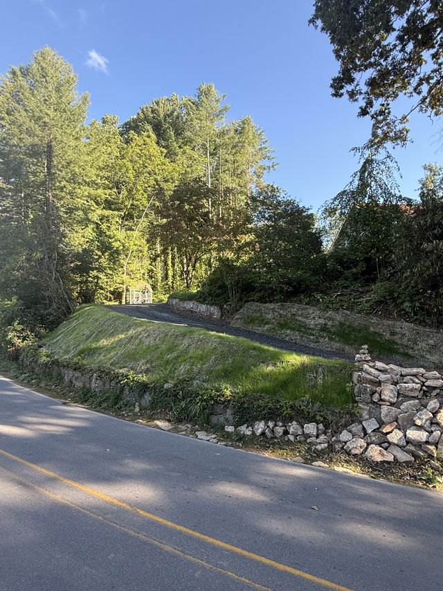 Gravel driveway entrance with a dry-stack stone retaining wall and graded grassy embankment along a wooded western North Carolina road