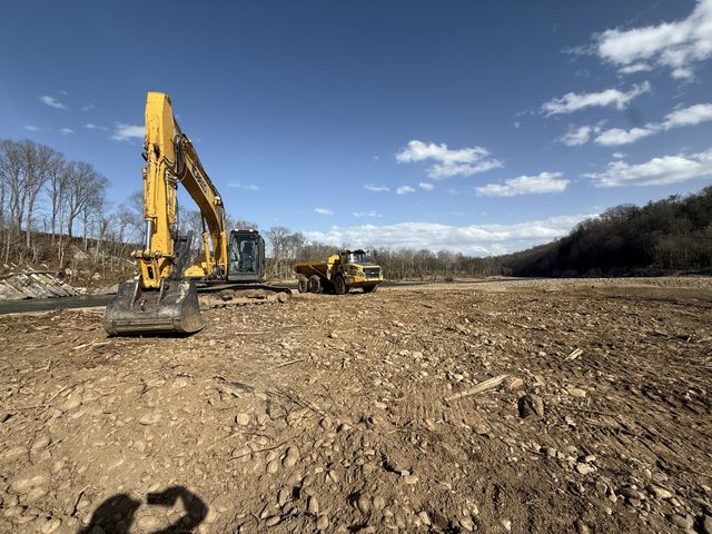 Kobelco excavator and articulated haul truck on a cleared river floodplain under a blue sky, western North Carolina