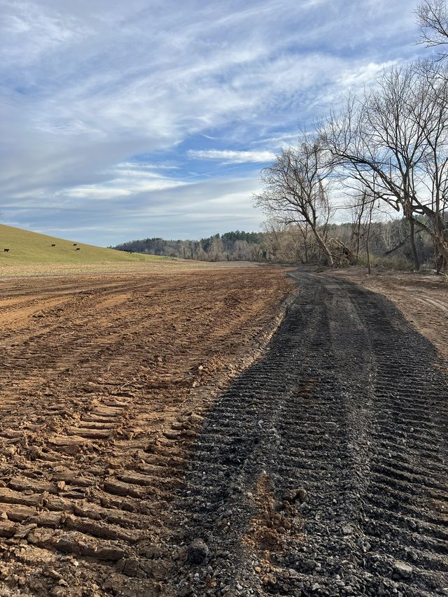 Freshly graded dirt meeting a newly laid gravel road cutting through rural pastureland, cattle grazing on a distant hillside