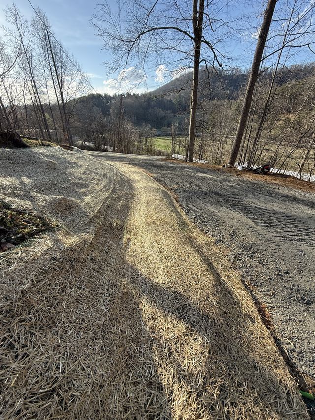 Newly graded gravel driveway curving through a wooded hillside with straw erosion matting on the embankment and a mountain valley beyond