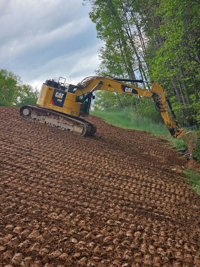 Tracked excavator shaping a graded slope on a steep wooded western North Carolina hillside, arm extended into the tree line