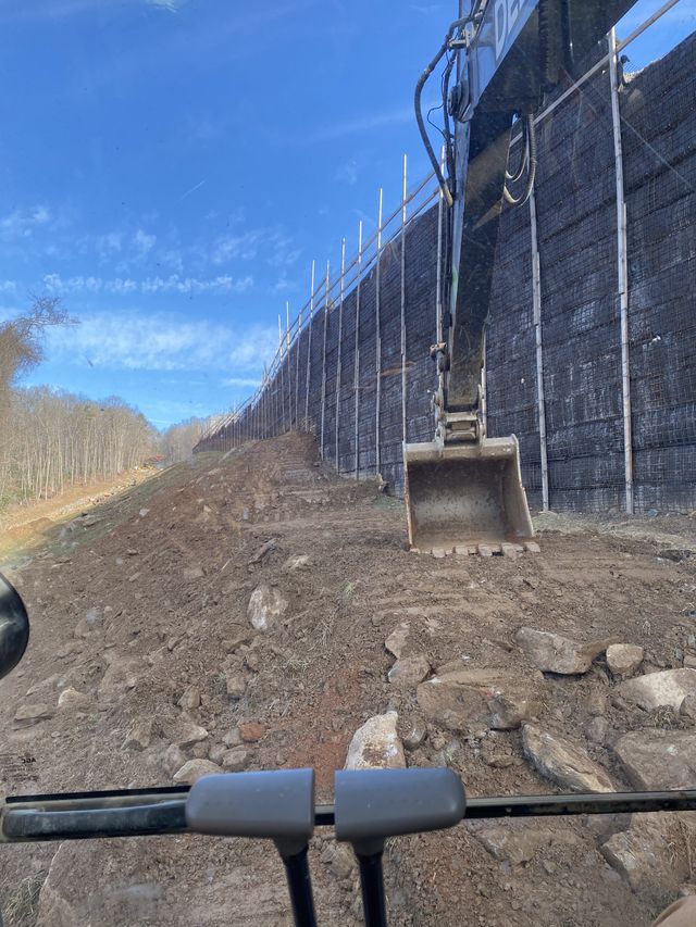 Operator's view of a wire-mesh reinforced retaining wall under construction along a wooded ridge under a clear blue sky