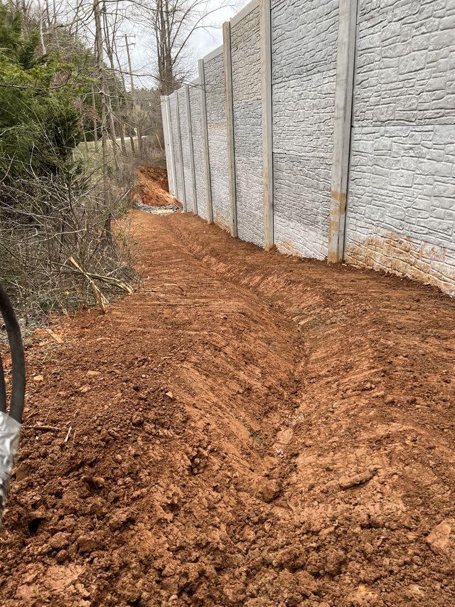 Long stone-pattern retaining wall running alongside a freshly graded red-clay path with strong receding perspective, western North Carolina