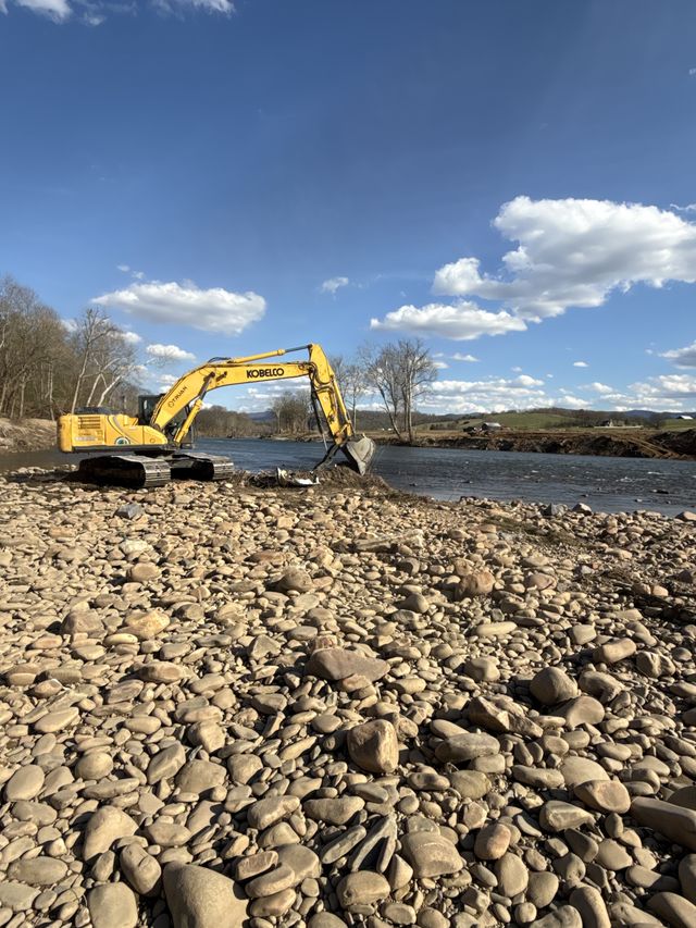 Kobelco excavator clearing flood debris at a river's edge with a rocky cobble bank and farmland in the distance, western North Carolina