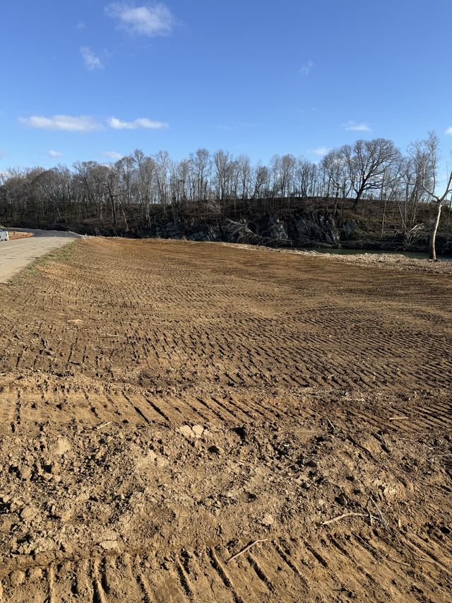 Wide finish-graded field running along a river with a rocky bluff and a gravel road alongside under a blue sky