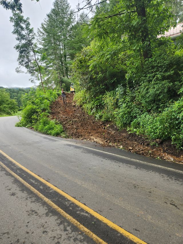 Cleared red-clay embankment along a curving two-lane mountain road in western North Carolina, brush and bank cut back to open up sightlines